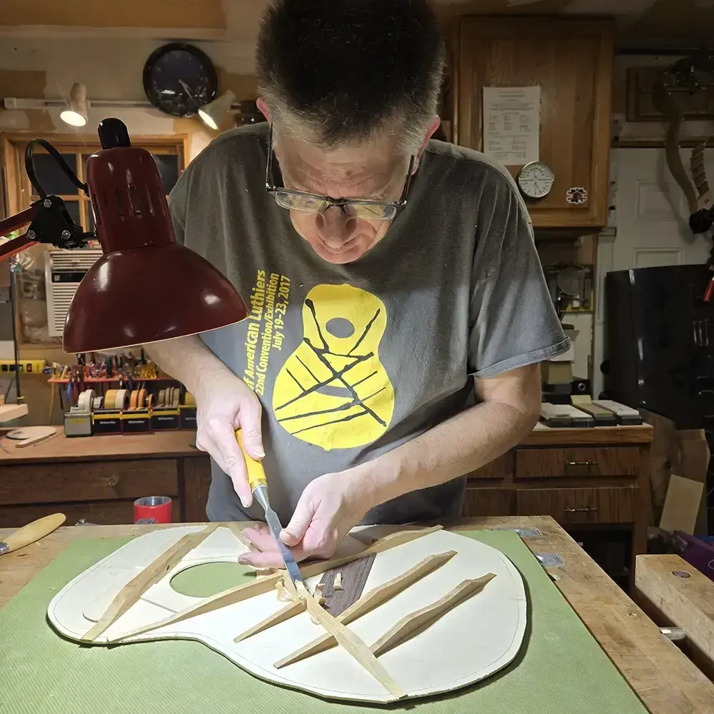 Luthier carving wooden guitar bracing by hand at a workbench in a workshop